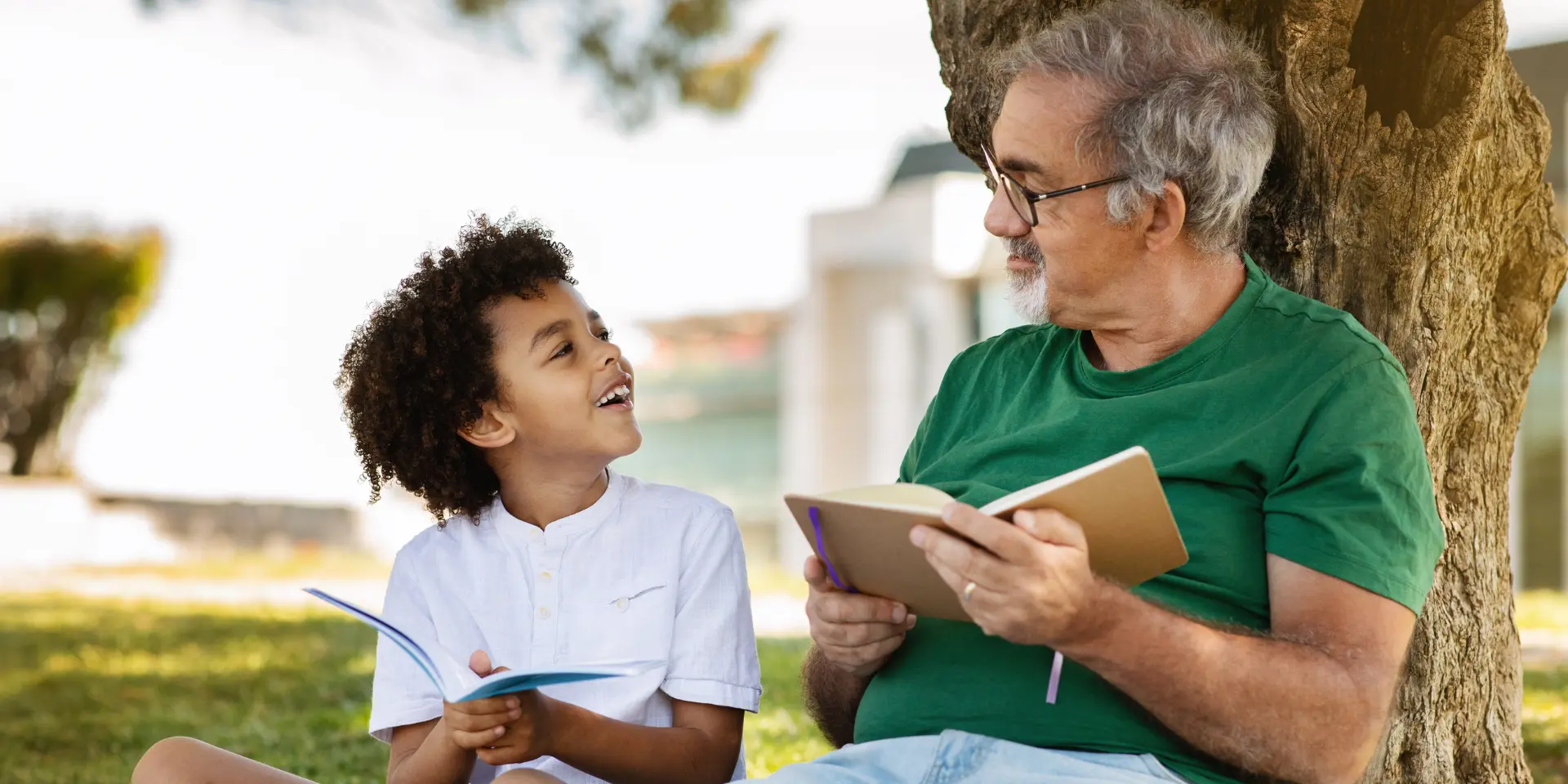 Glad senior man with beard and black little boy sit on plaid read a book relax in park