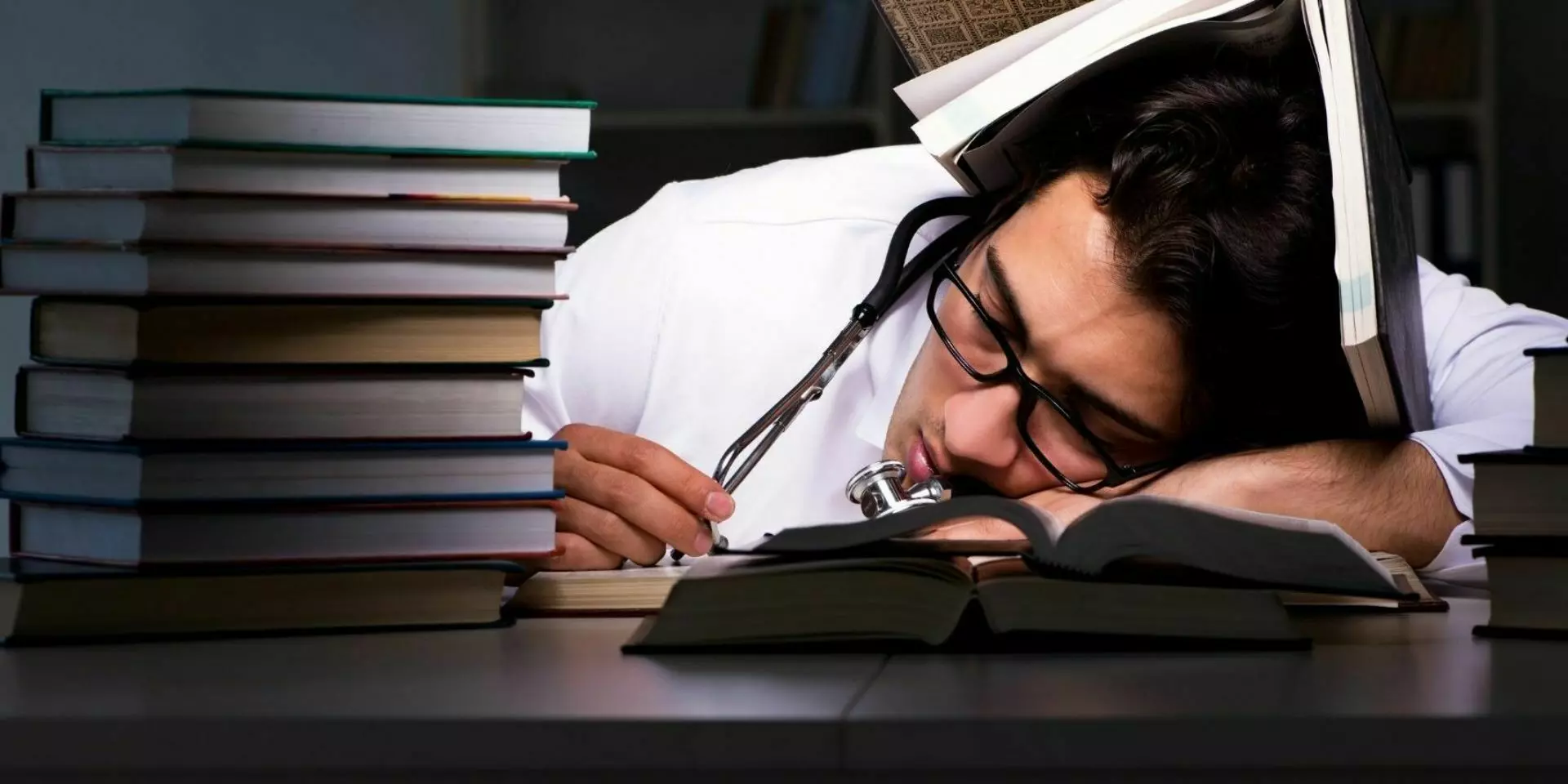 a doctor asleep at a desk surrounded by books with one over his head