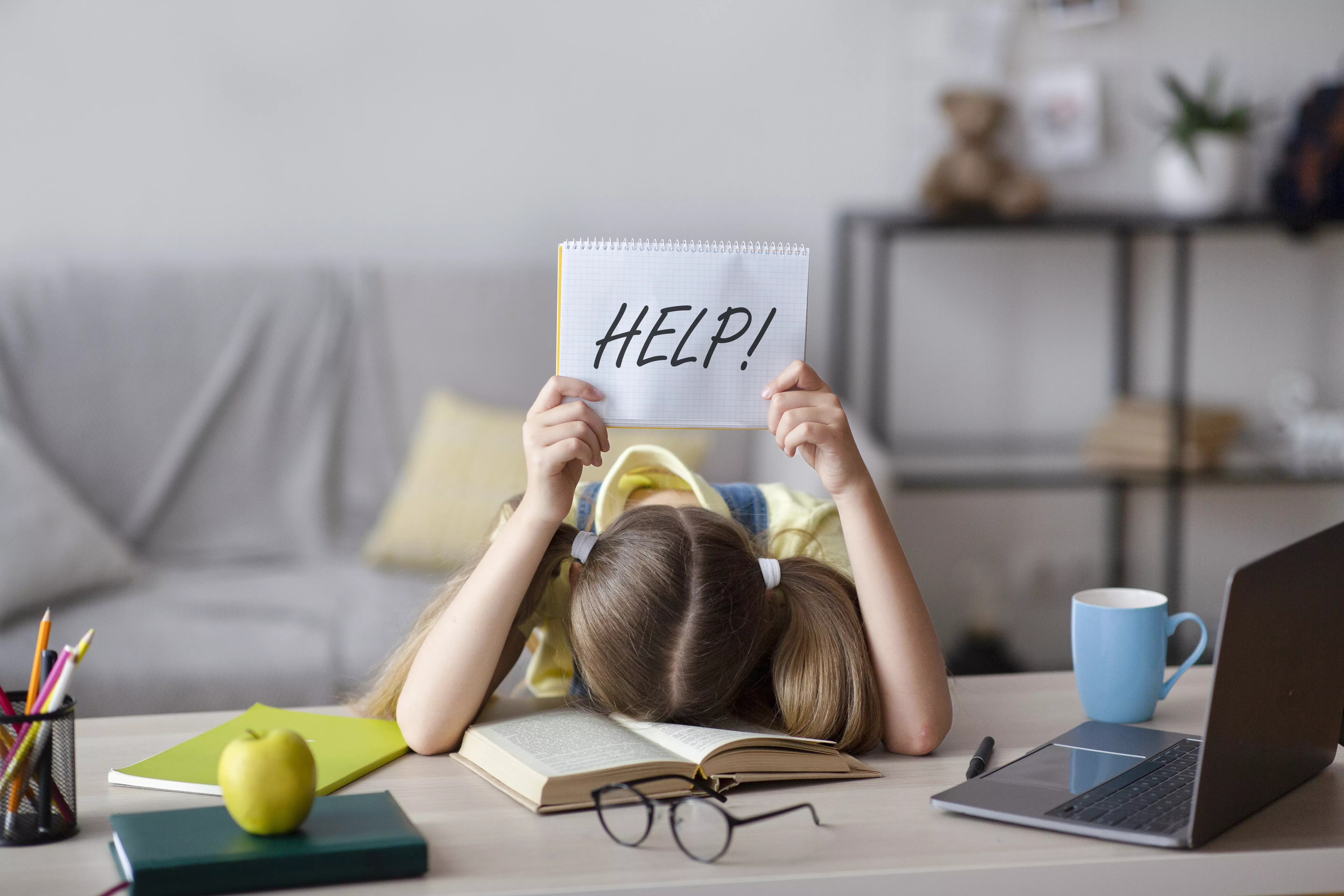 a frustrated girl with her head on the desk in front of her, holding up a help sign