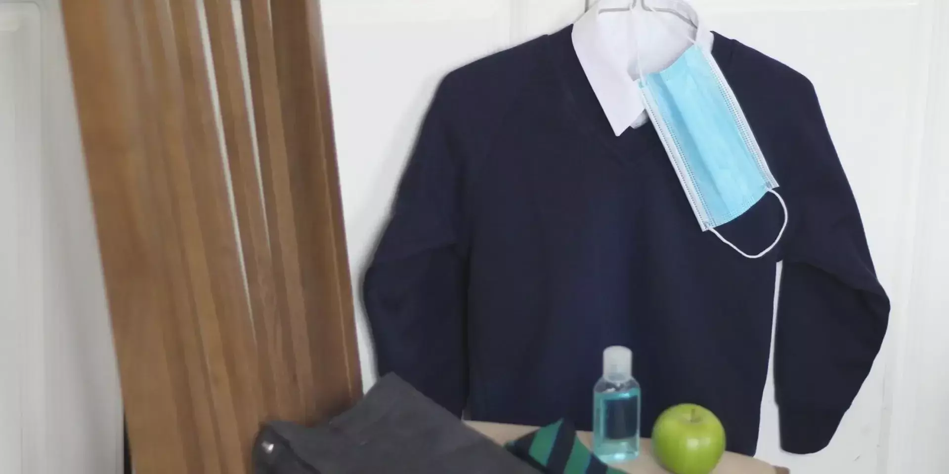 a school jumper hung up with a face mask in the collar and an orange and a bottle of hand sanitizer below it