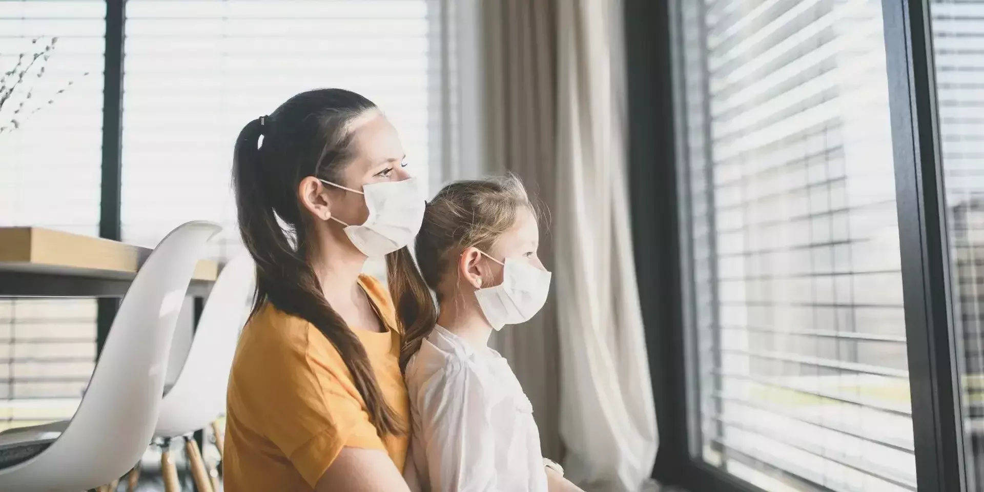a mother holding her daughter on her lap both wearing facemasks and looking out the window